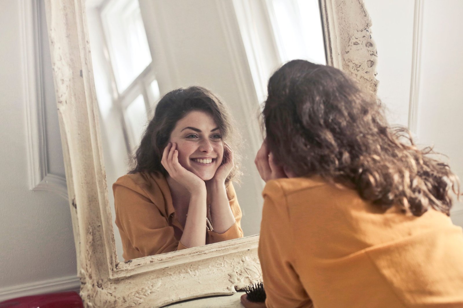 Home A cheerful woman smiles at her reflection in a vintage-style mirror, exuding positivity and warmth.