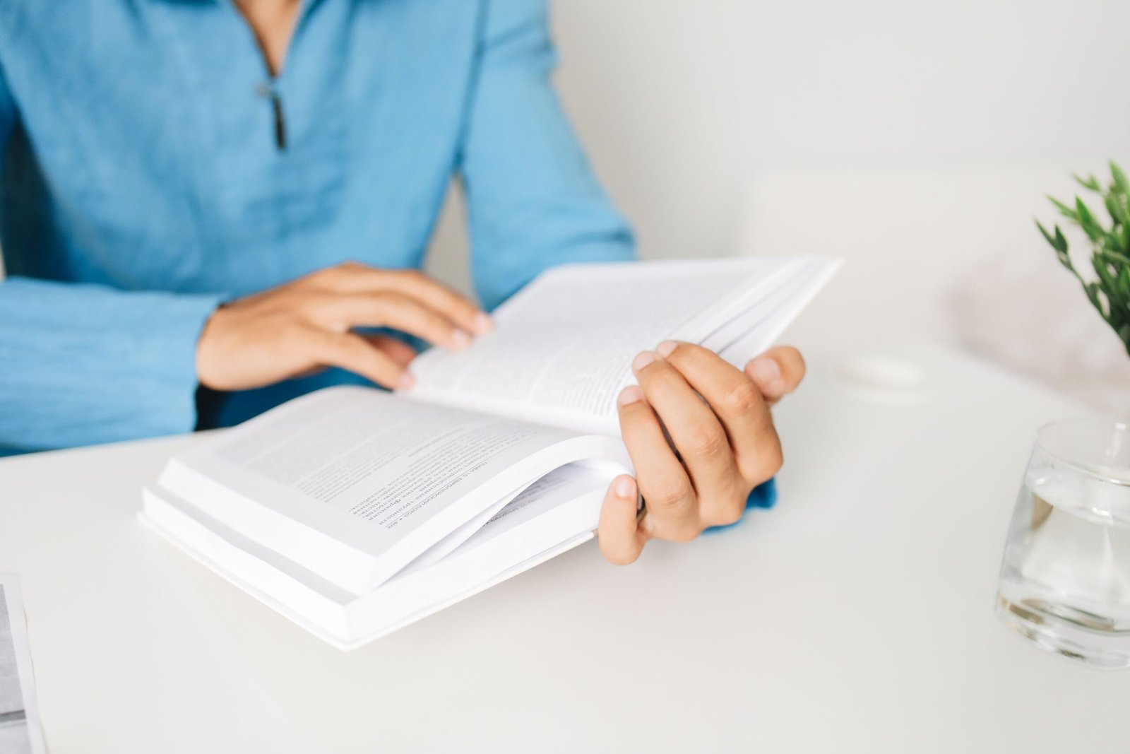 Home Person in a blue shirt studying a book at a white desk indoors with a plant.